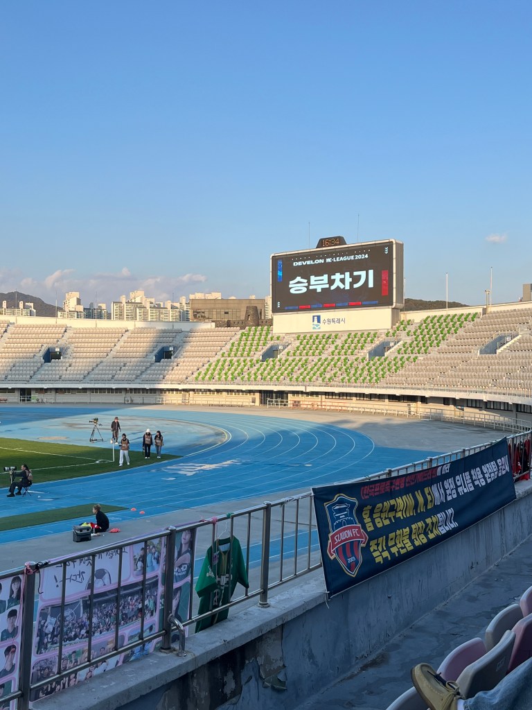 Inside an open air bowl-shaped stadium with a blue running track, a giant screen indicates that a penalty shootout is about to take place