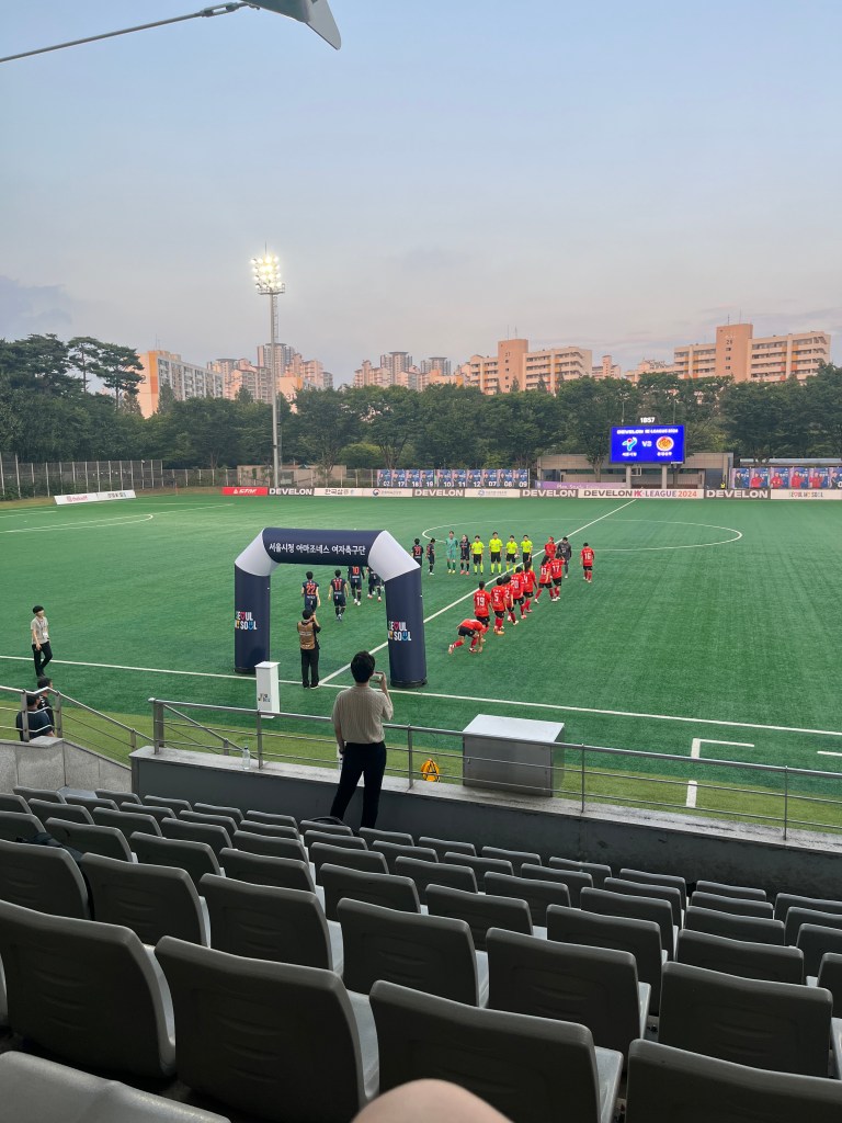 Players walk on to an artificial turf football pitch. There is an inflatable archway they walk through as they line up, and a big screen in the background
