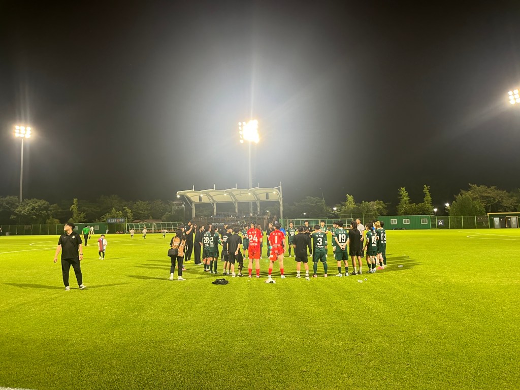 Football players in green uniforms stand in a circle along with their coaches on an artificial pitch under floodlights