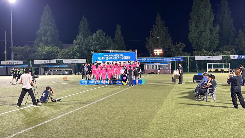A women's football team dressed in pink uniforms receiving a abanner and trophy on a small temporary stage area on an artificial football pitch.