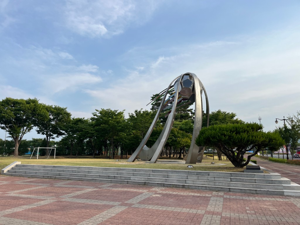 Next to a paved area, a large metal statue of a football, held up between large sweeping metal supports, with shards emerging from it as if in motion.