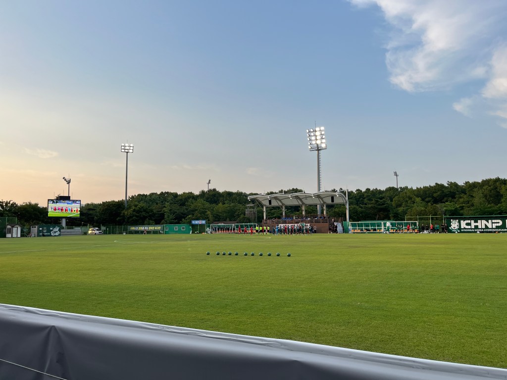 A football pitch before a match. In the foreground is a row of footballs ready for a giveaway. On the far side of the pitch two teams are lining up and giving each other high fives.