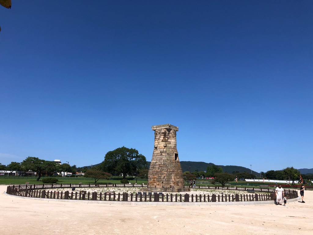 An ancient Korean astronomical observatory built of stone. It is around 9 metres high with a cylindrical body standing on a base, and with a square top. There is a small window halfway up. In the background are green mountains and trees, and a bright blue sky.