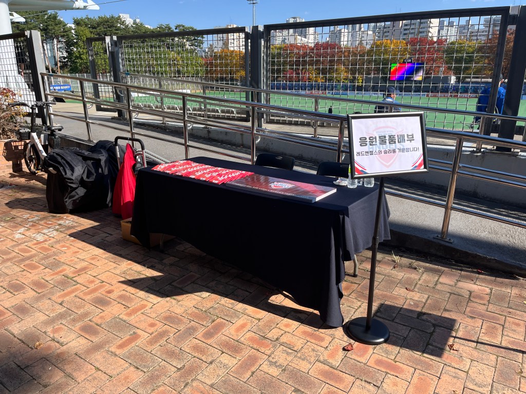 Outside the entrance to a small football ground, there is a table with small banners and foldable clappers marked with the logo of Incheon Hyundai Steel Red Angels.