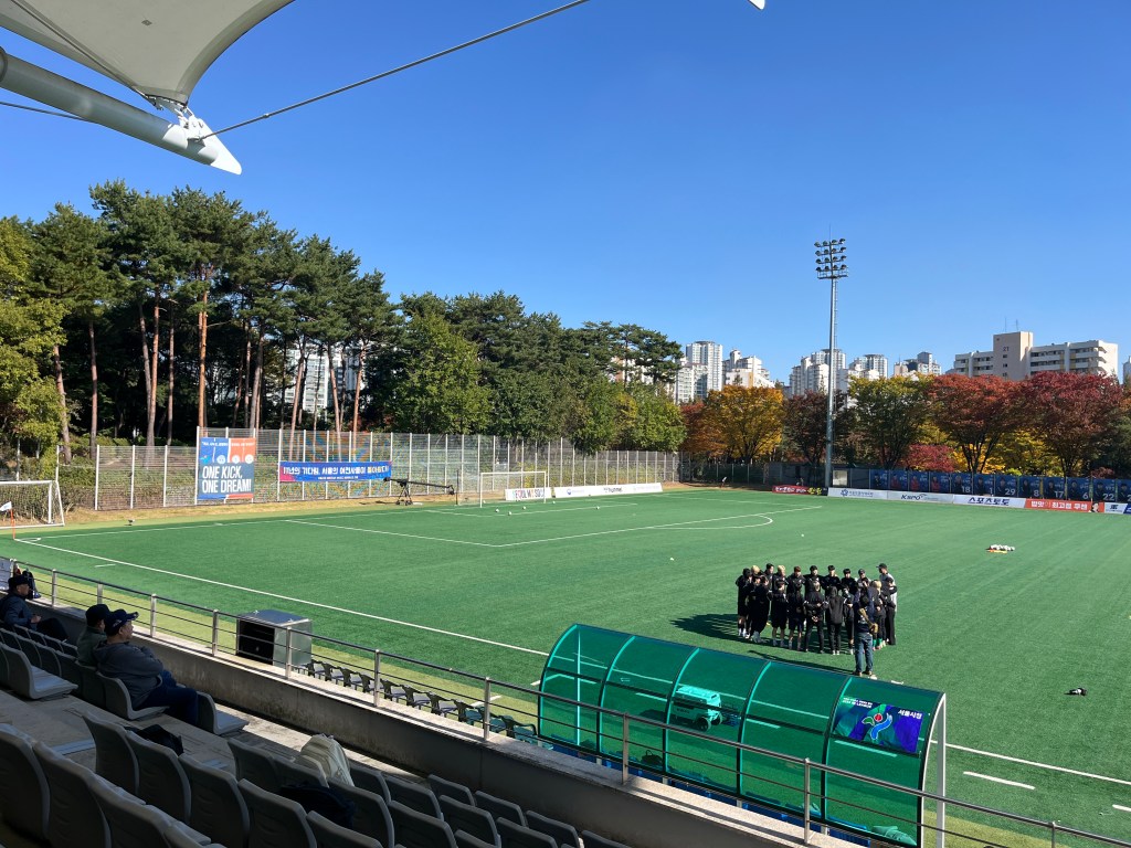 On an artificial football pitch surrounded by autumnal trees and with a bright blue sky overhead, a women's football team and their coaches huddle together in front of their dugout.