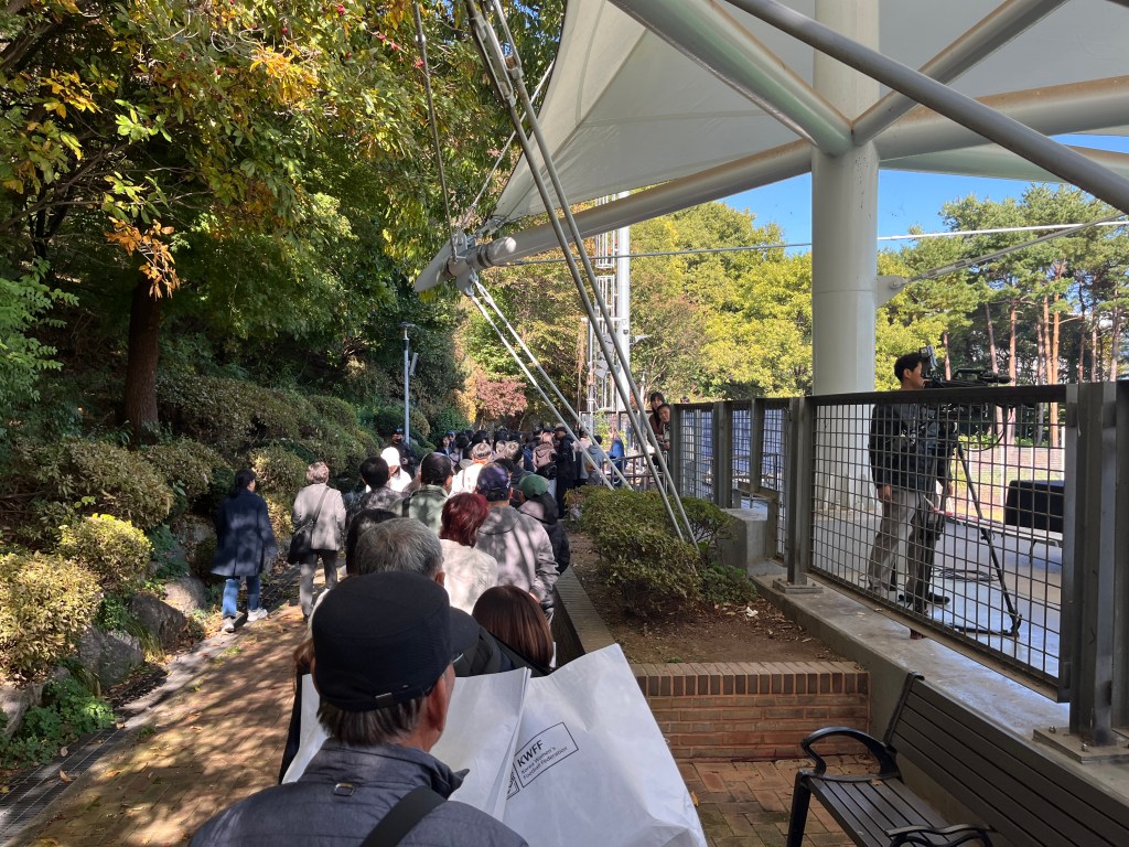 People stand in a queue outside a small football ground. Some of them are holding bags marked 'Korea Women's Football Federation'.